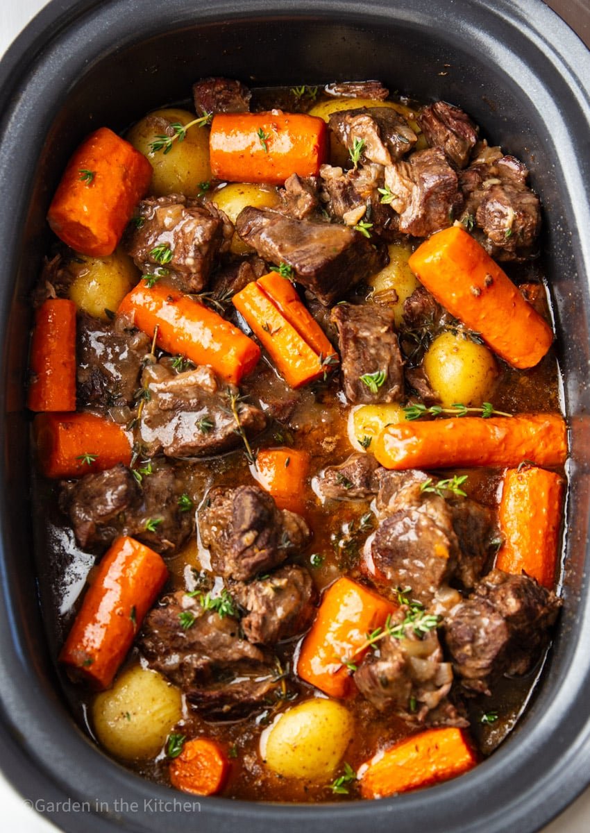 Slow cooker beef stew with chunks of beef, carrots, baby potatoes, thyme in a black slow cooker.