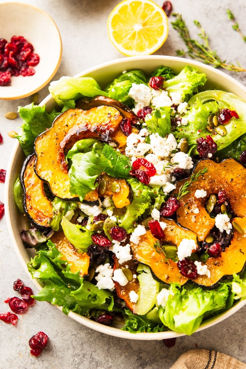 Fall salad with roasted squash, salad greens, dried cranberries, goat cheese and pumpkin seed in a white bowl with dried cranberries on a plate, lemon, pumpkin seeds and rosemary.