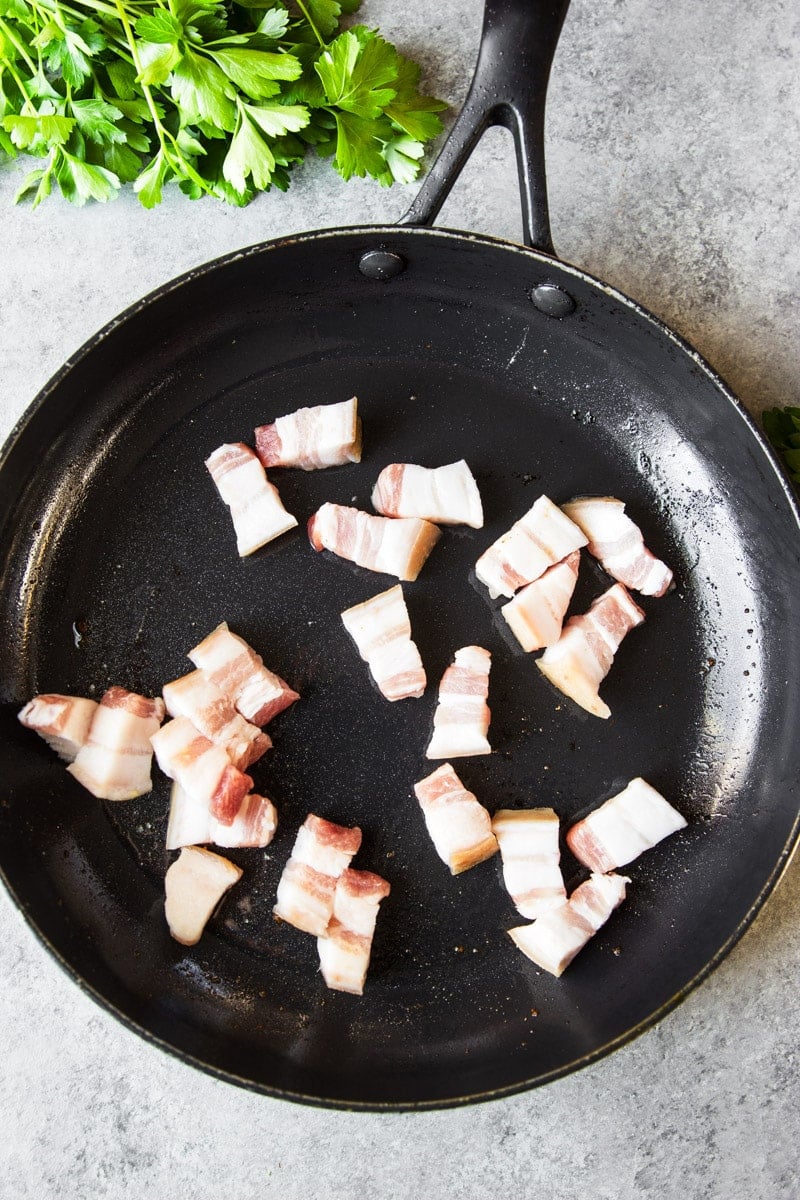 Chunks of bacon being fried in a black cast iron pan with parsley. 