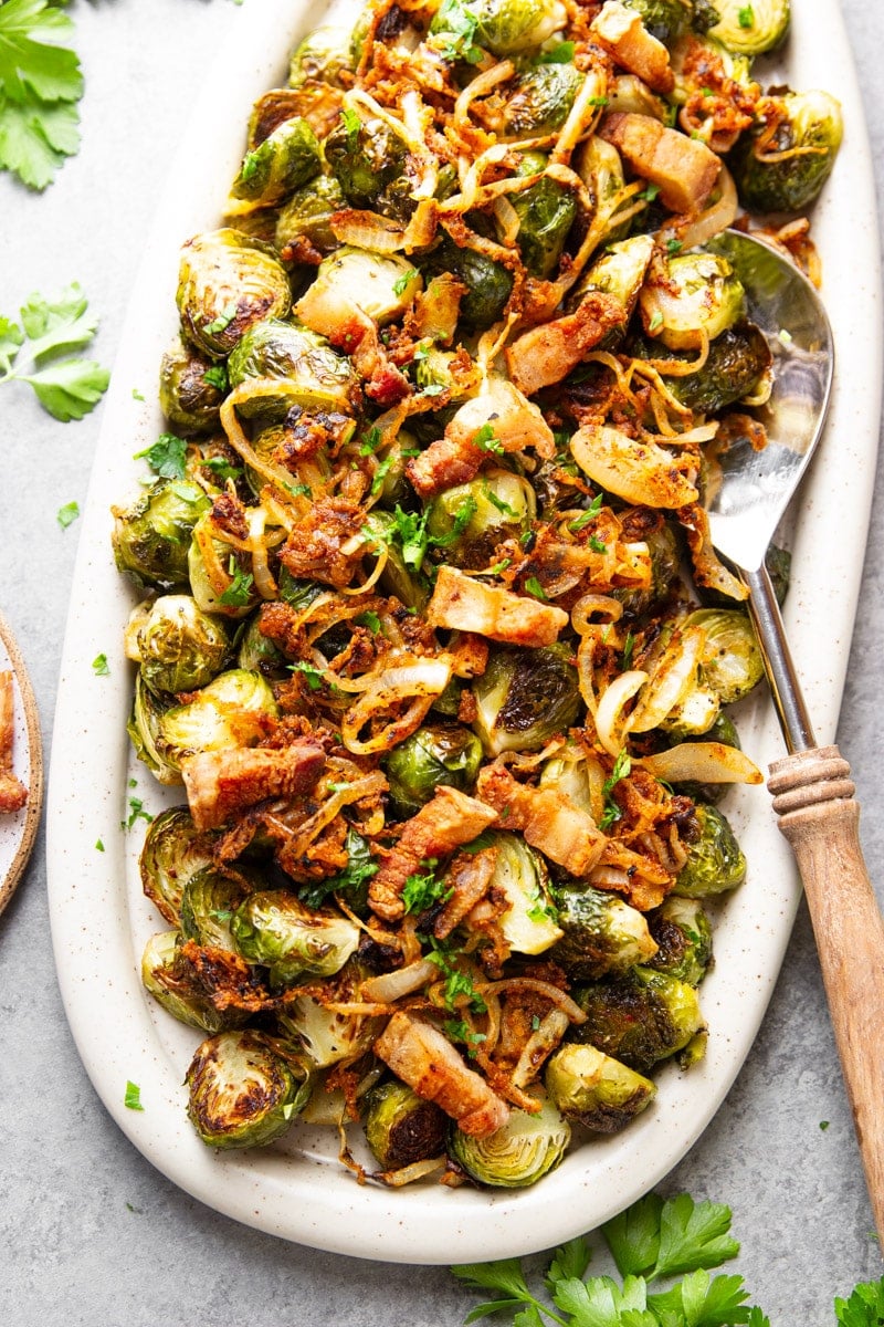 Roasted Brussels sprouts with crispy onions and bacon in a white serving dish with a stainless steel spatula with a wooden handle and parsley.