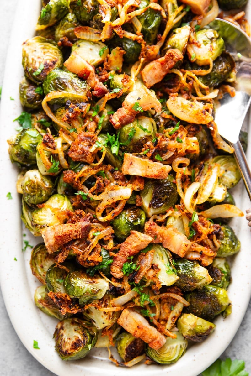 Roasted Brussels sprouts with crispy onions and bacon in a white serving dish with a stainless steel spatula. 