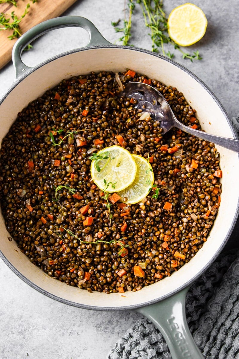 Cooked French lentils with carrots, celery, thyme, lemon slices, silver spoon in a Le Creuset pot, lemon, wooden cutting board.