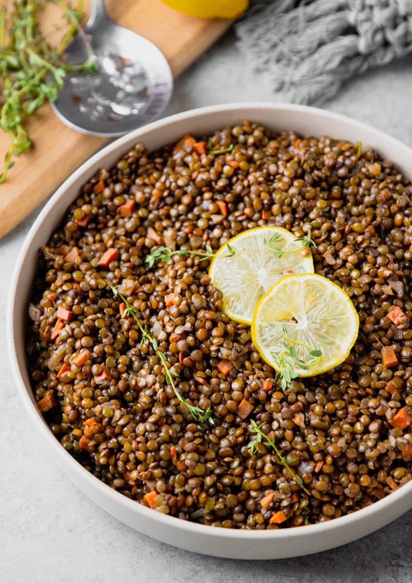 A pot of French lentils with lemon slices and thyme, wooden cutting board silver spoon, lemon and grey scarf. 