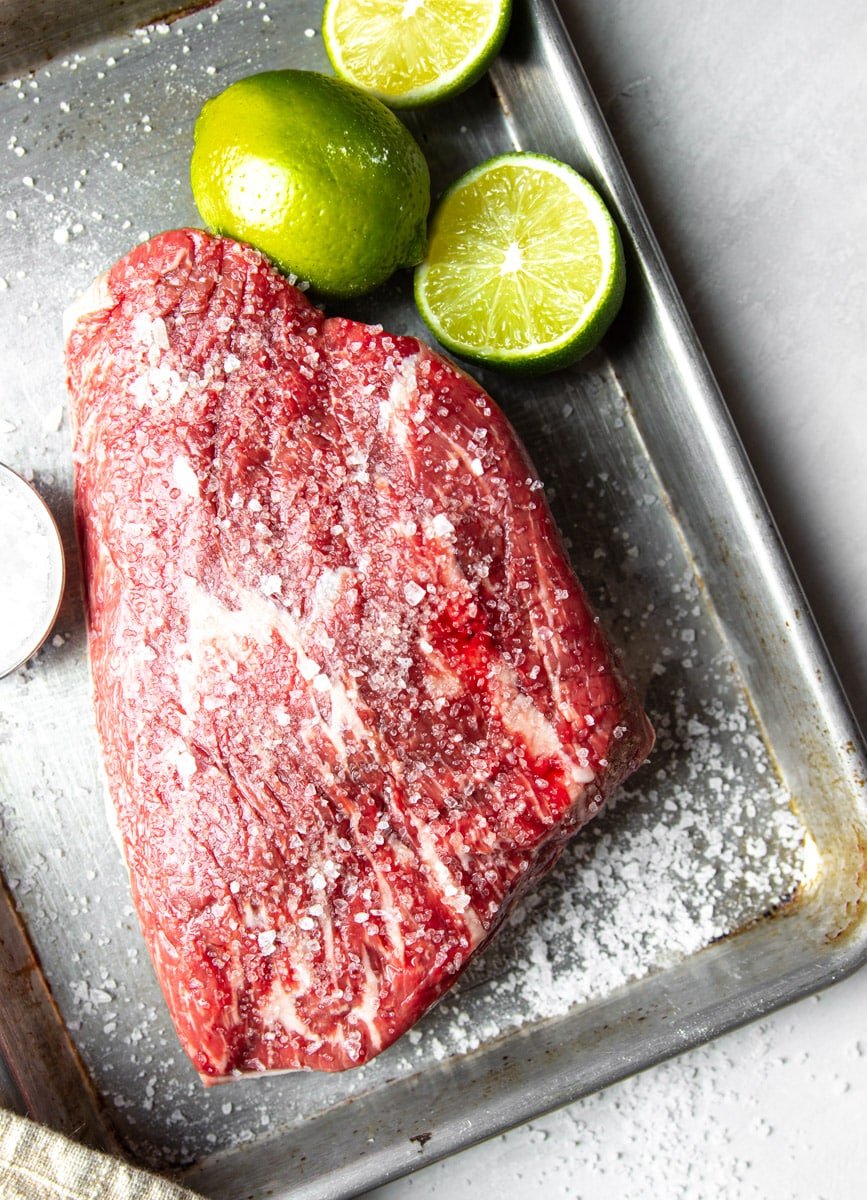 Raw picanha steak seasoned with salt and limes on a stainless steel baking sheet. 