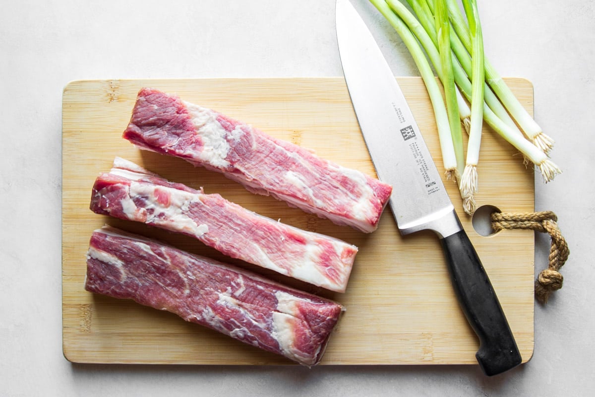 Strips of pork belly on a wooden cutting board with a knife and green onions. 