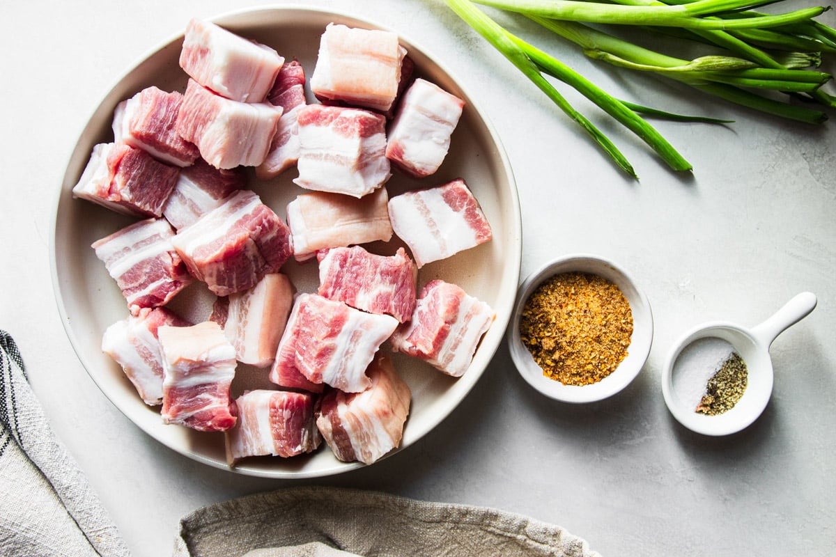 Cubed pork belly in a white bowl with the seasoning rub and green onions. 
