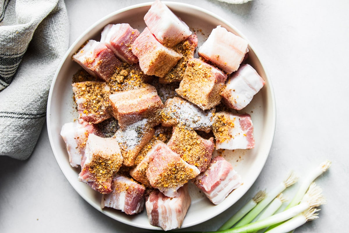 Diced pork belly in a white bowl with the seasoning rub, green onions and a white and black dish towel. 