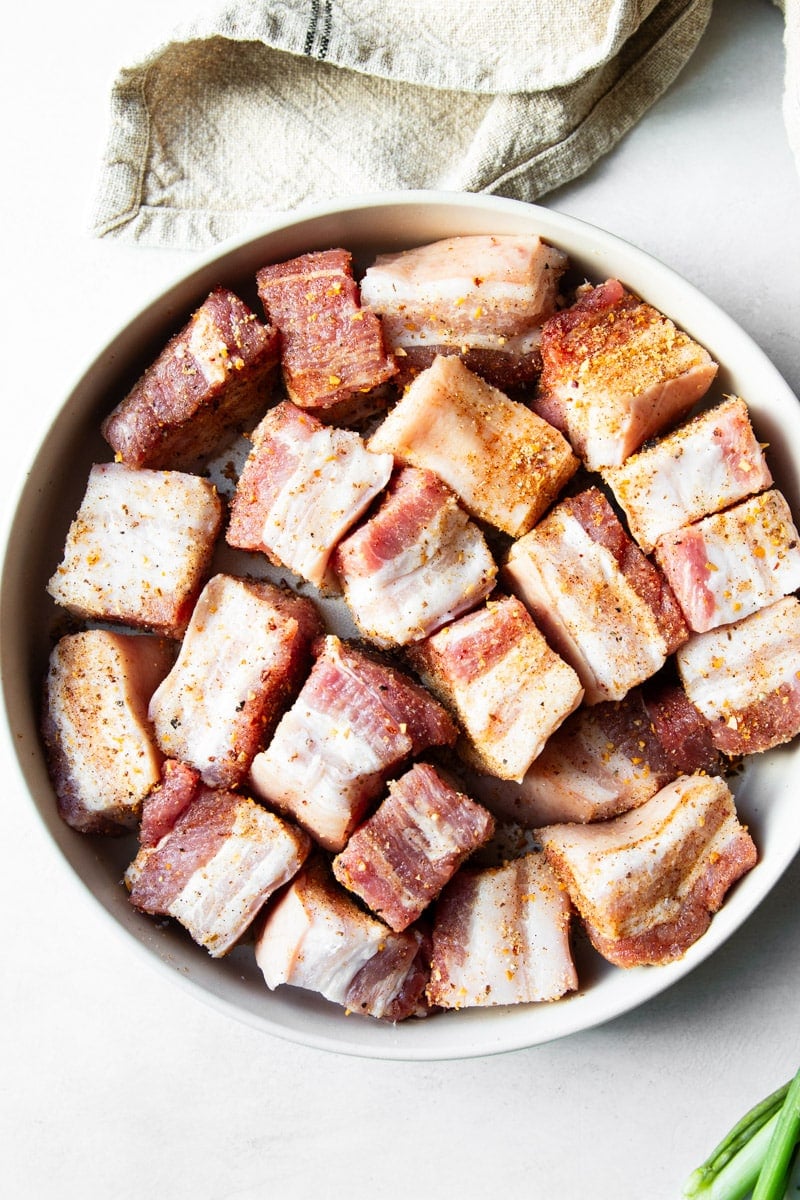 Diced pork belly with a seasoning rub in a white bowl, and green onions. 