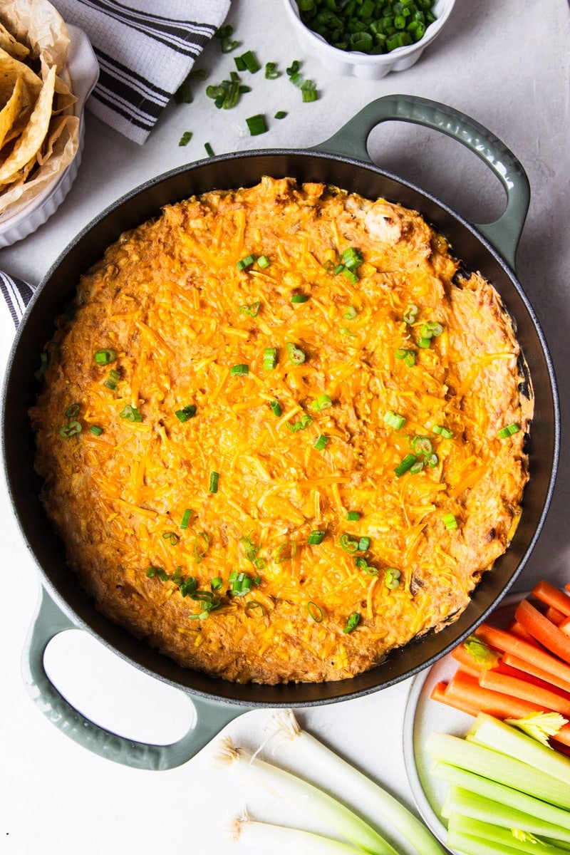 Baked buffalo chicken dip in a skillet, green onions, white dish towel, corn chips, celery, and carrots. 