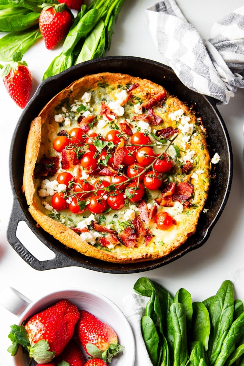 Baked Dutch baby in a cast iron skillet with cherry tomatoes, loose strawberries, strawberries in a white bowl, spinach wrapped in a dish towel. 