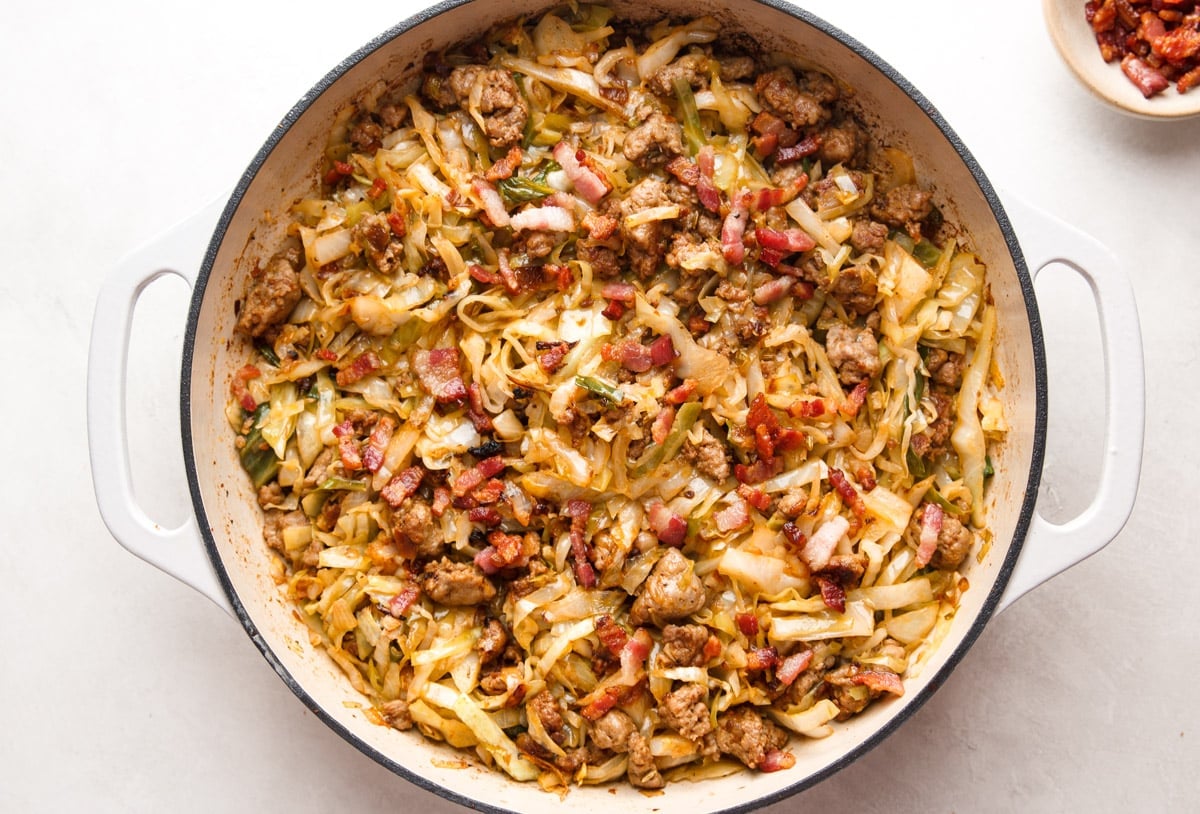 Fried cabbage with sausage in a white skillet, bowl of chopped bacon. 