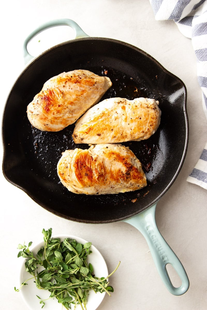 Seared chicken in a cast iron skillet, fresh oregano in a white bowl and a blue and white dish towel. 