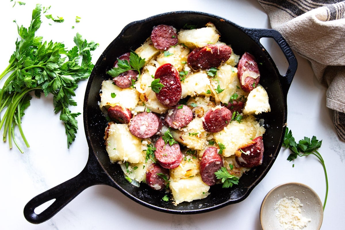 Fried yuca and kielbasa in a cast iron skillet, parsley, parmesan cheese, and a white and grey dish towel. 