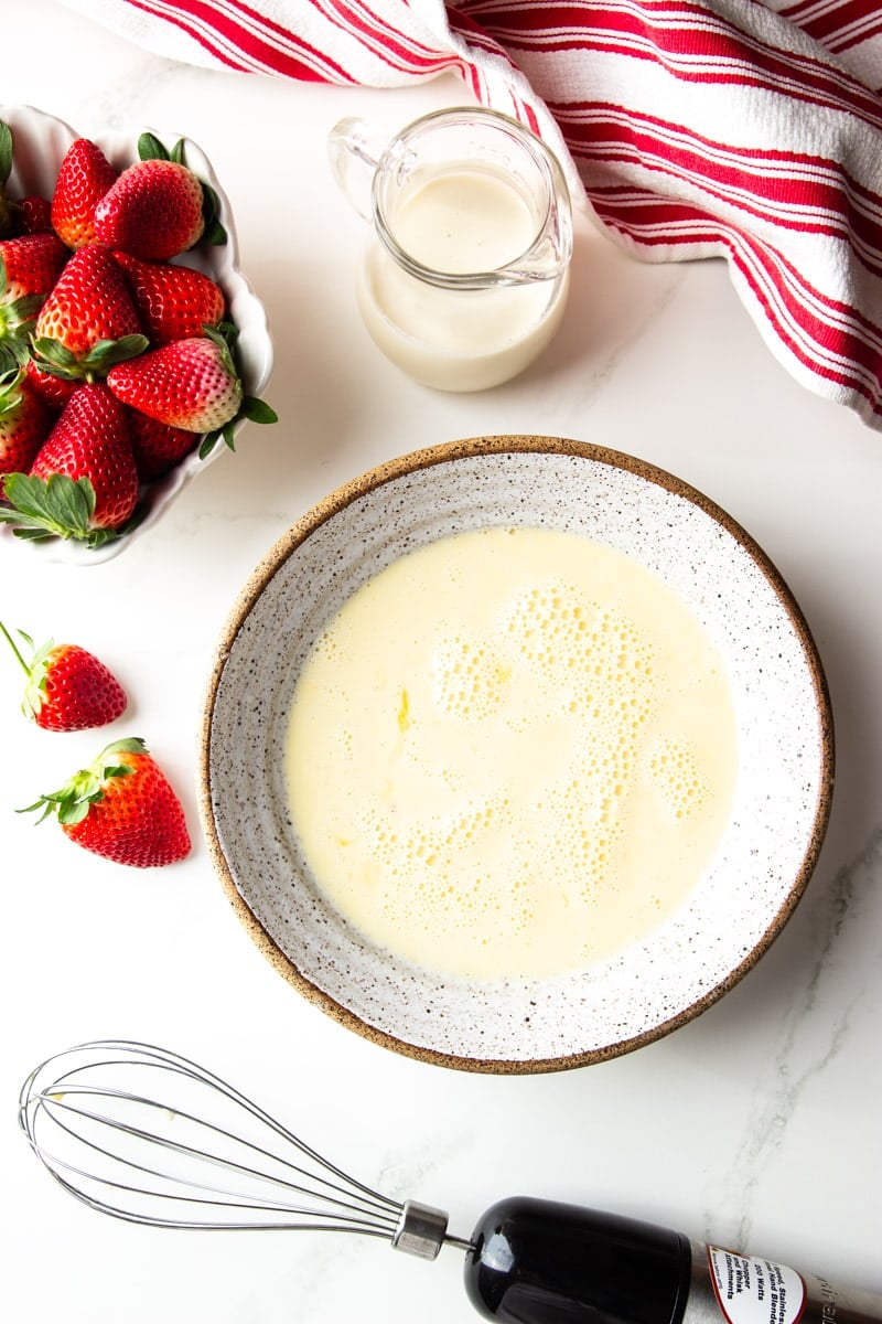 Strawberries, whisked eggs in a bowl, heavy cream, electric whisk, white and red dish towel. 