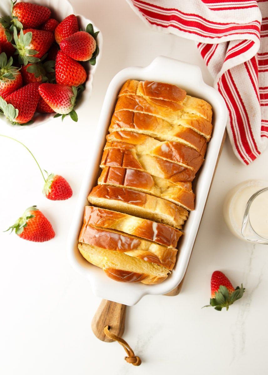 Brioche bread in a white casserole dish, strawberries, cream, and a white and red dish towel. 