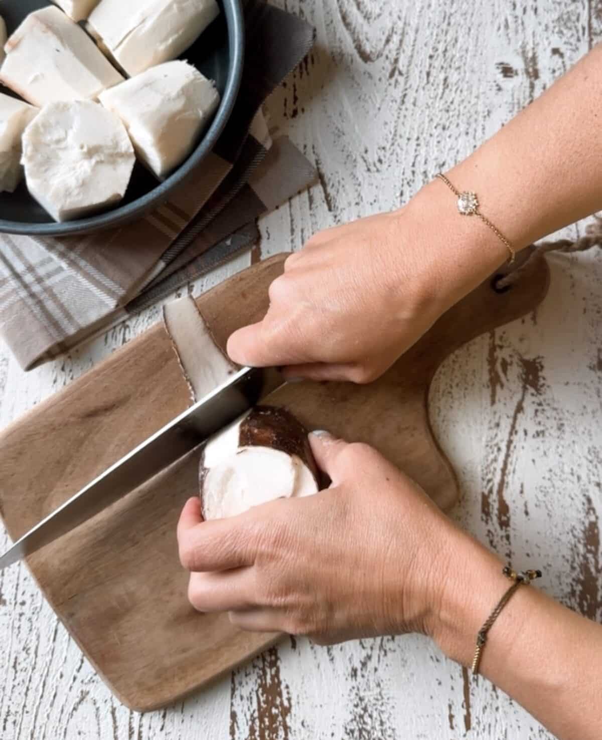 Peeling yuca on a wooden cutting board with yuca in a bowl on a white and brown dish towel. 