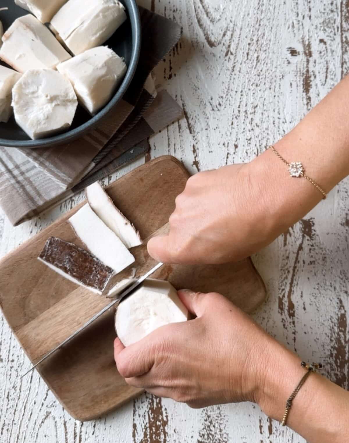 Peeling yuca on a wooden cutting board with yuca in a bowl on a white and brown dish towel. 