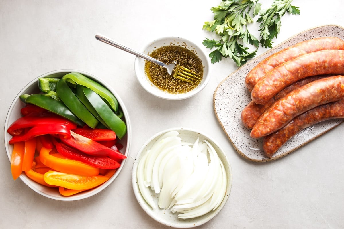 Ingredients measured for sheet pan sausage and peppers recipe. 5 Italian sausages, green, red and orange bell peppers, sliced onion and a small bowl of marinate
