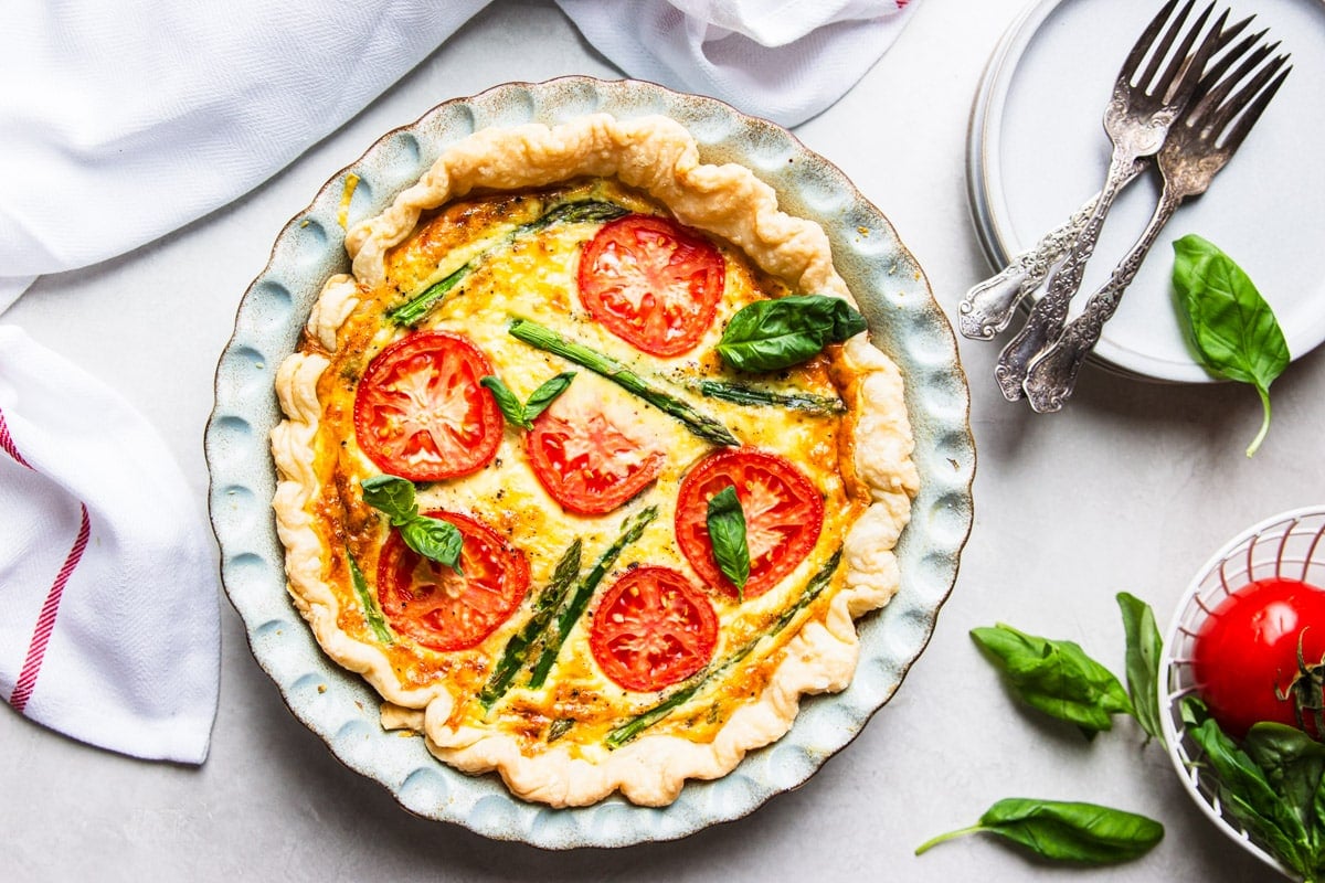 Baked tomato and asparagus quiche, fresh basil, tomato, white and red dish towel, forks and a white plate. 