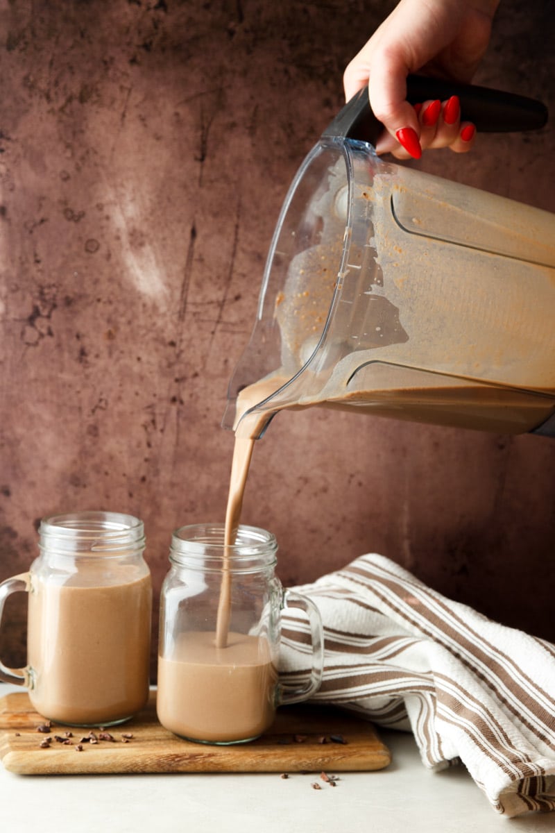 Chocolate Coffee Protein Smoothie being poured into a glass jar on a wooden cutting board, white and brown dish towel. 