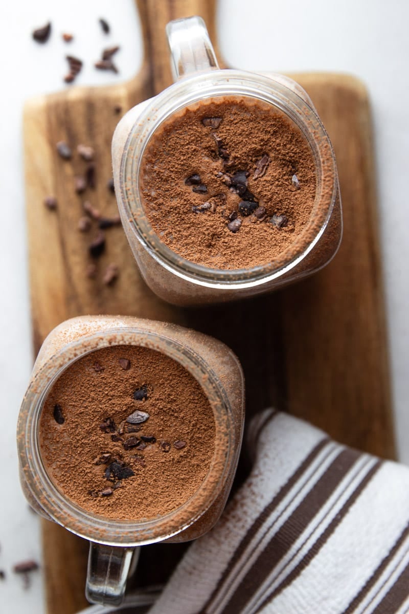 Chocolate Coffee Protein Smoothie in a glass jar with chocolate powder and cocoa nibs on top, on a wooden cutting board and a white and brown dish towel. 