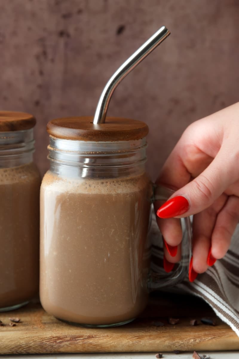 Chocolate coffee protein smoothie, stainless steel straw, wooden cutting board, chopped chocolate and a white and brown dish towel. 