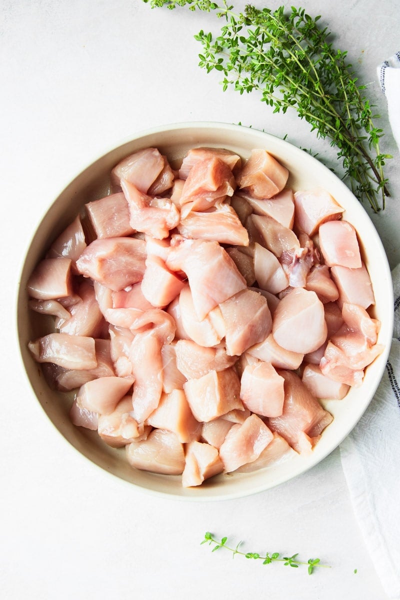 Raw cubed chicken in a white bowl, fresh thyme and a black and white dish towel. 