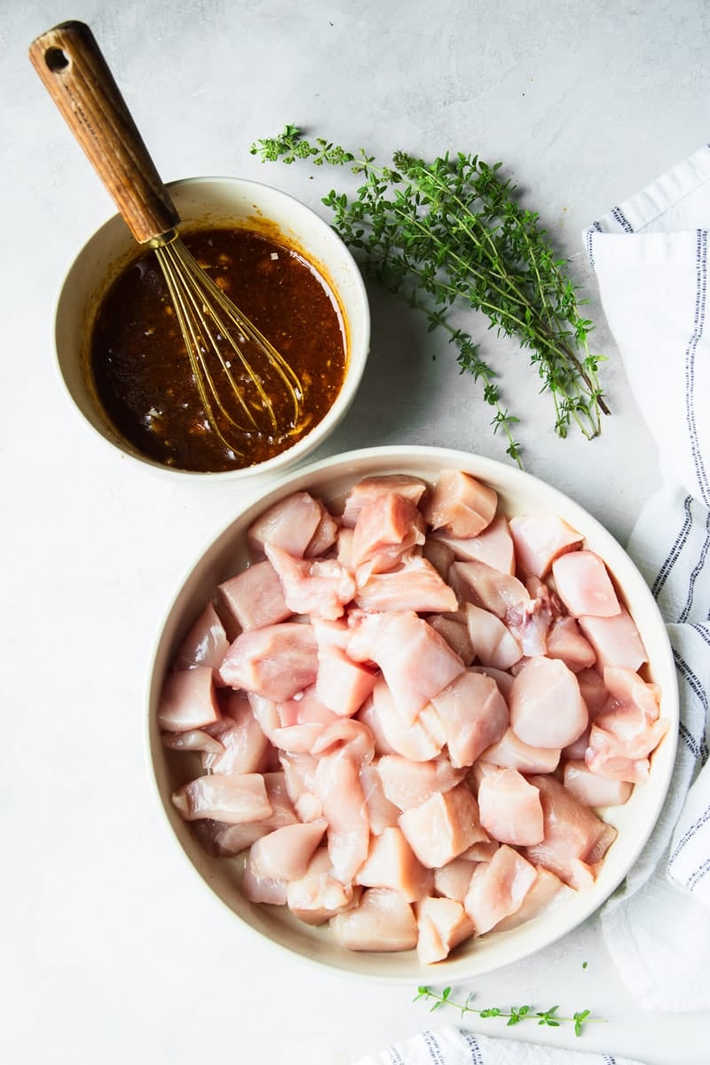 Fresh thyme, honey mustard sauce in a bowl with a whisk, raw chicken cubed in a white bowl and a black and white dish towel. 