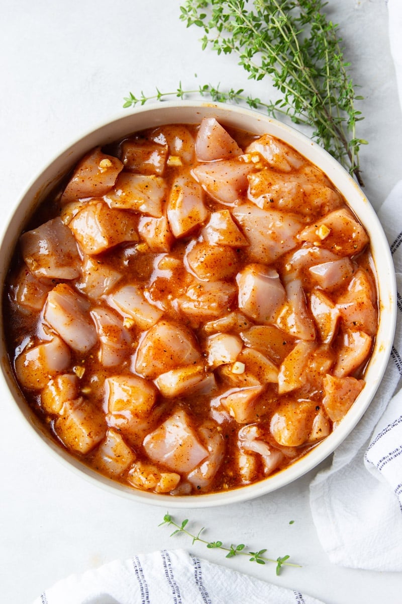 Cubed chicken marinating in a honey Dijon sauce in a white bowl, fresh thyme and a white and black dish towel. 