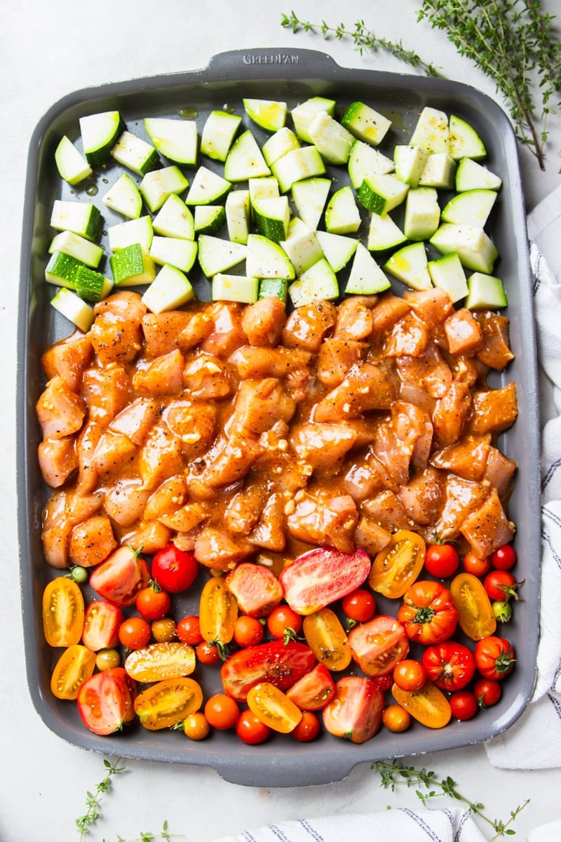 Cubed zucchini, marinated chicken cubes and sliced tomatoes on a baking sheet, white and black dish towel, fresh thyme. 