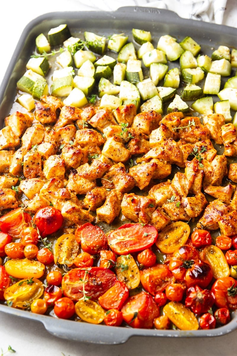 Roasted zucchini and tomatoes with cooked honey mustard chicken on a cookie sheet and a black and white dish towel.