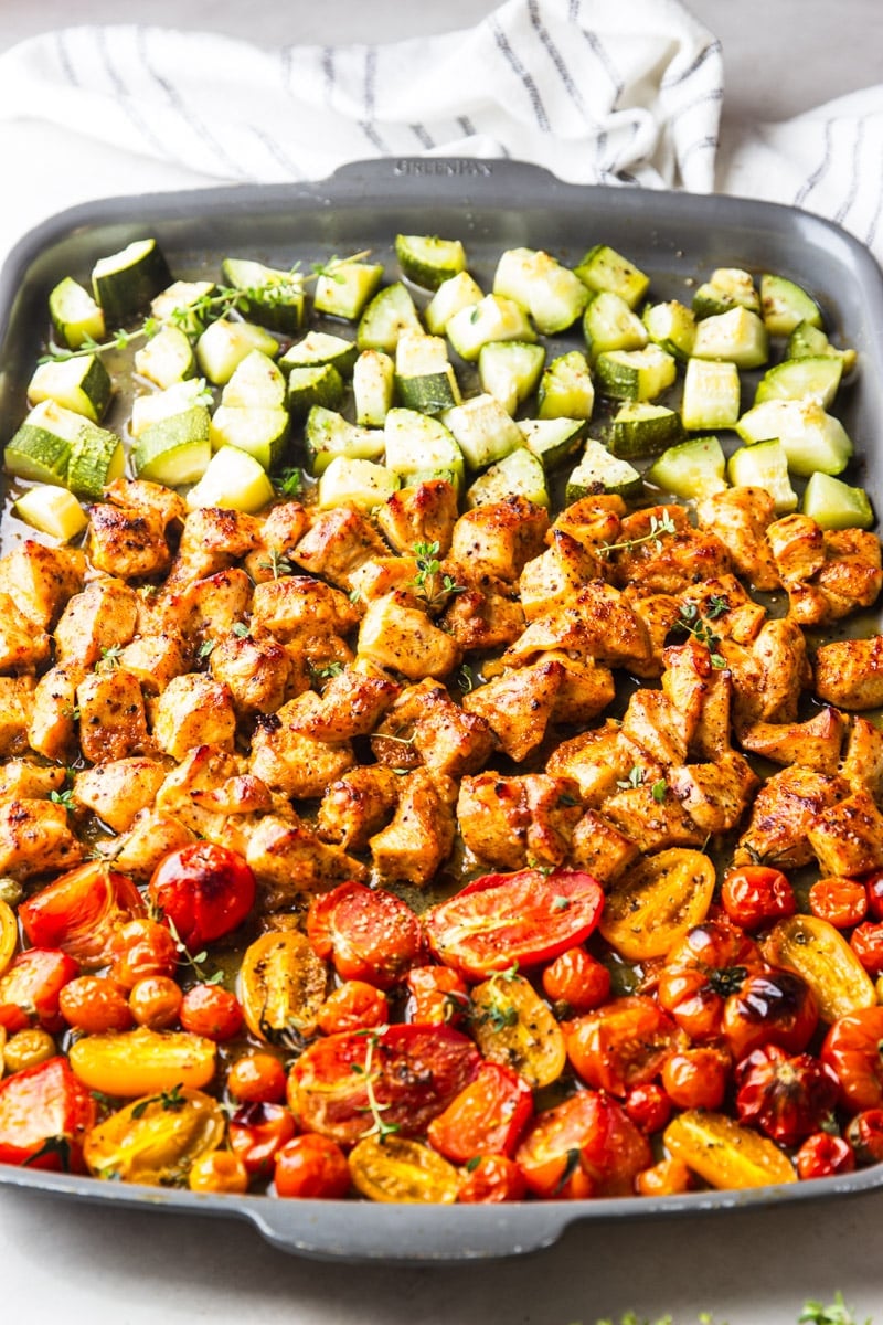 Roasted zucchini and tomatoes with cooked honey mustard chicken on a cookie sheet and a black and white dish towel.