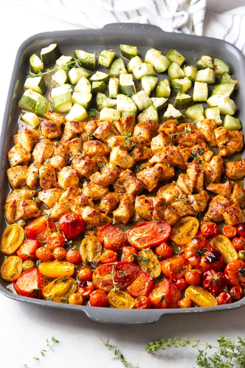 Sheet pan honey mustard chicken with zucchini and tomatoes on a baking sheet. Black and white dish towel and thyme. 