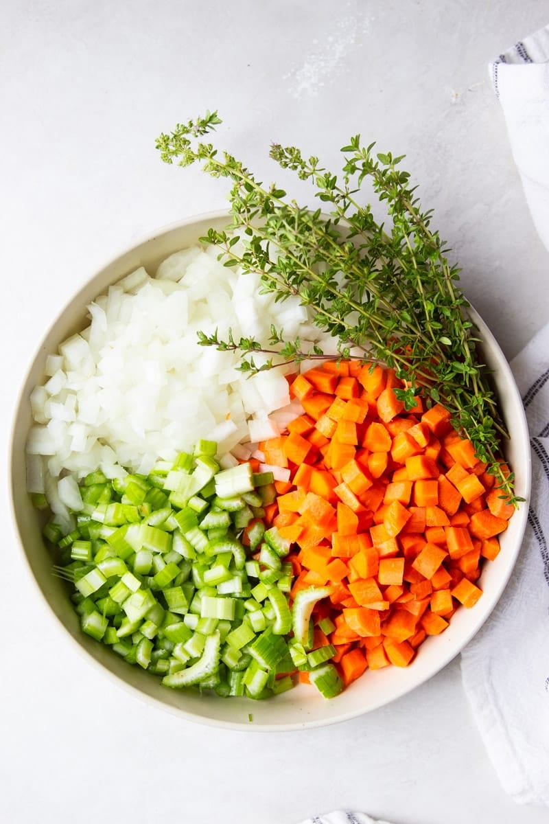 Chopped onion, diced carrots and celery, fresh thyme in a white bowl with a white and black dish towel.