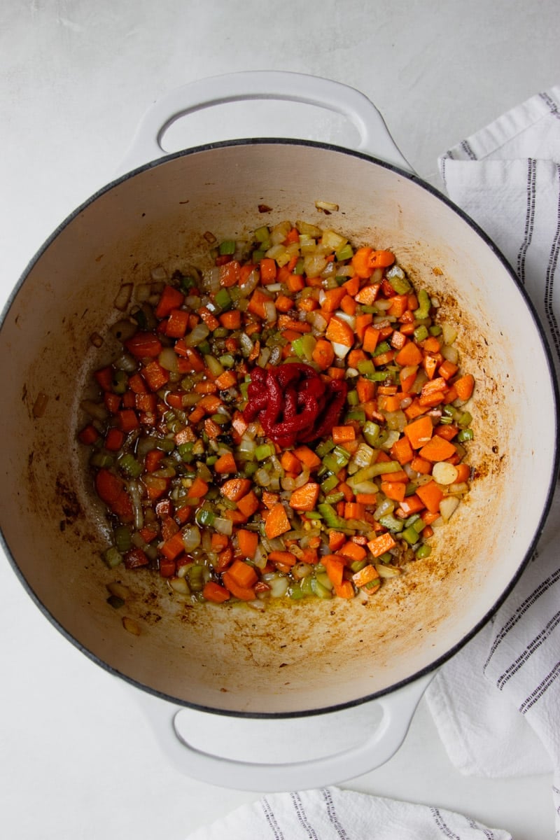 Sauteed veggies in a pot with tomato paste added. White and black dish towel. 