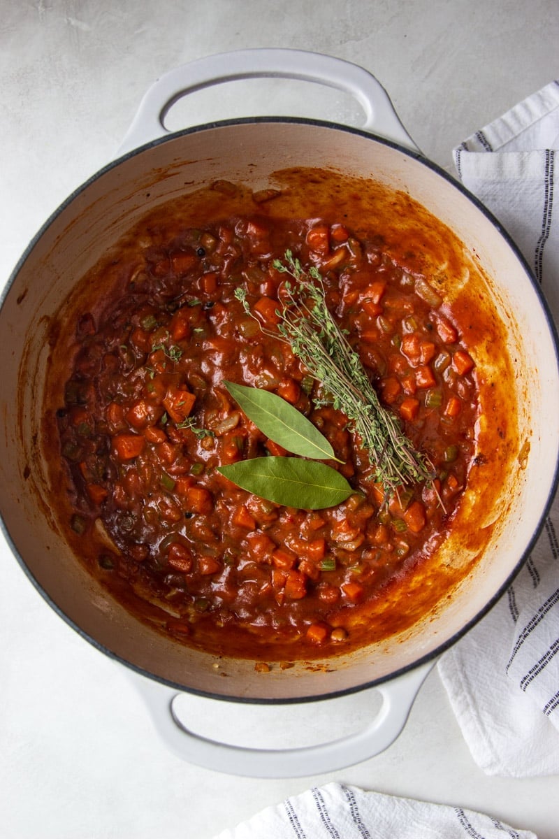 Sauteed veggies in a pot with fresh thyme and bay leaves and a white and black dish towel.