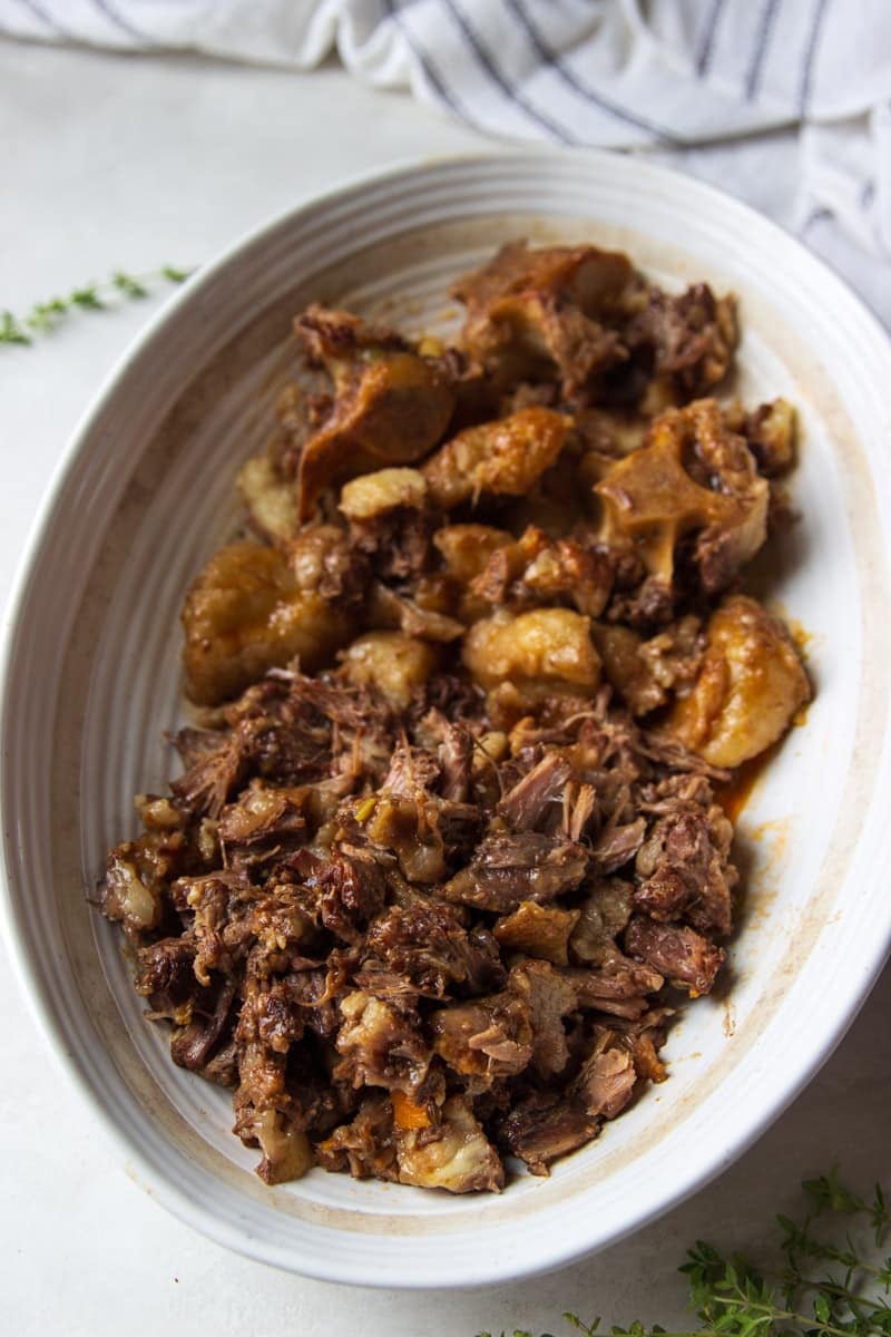 Cooked oxtail in a white bowl, fresh thyme and a white and black dish towel.