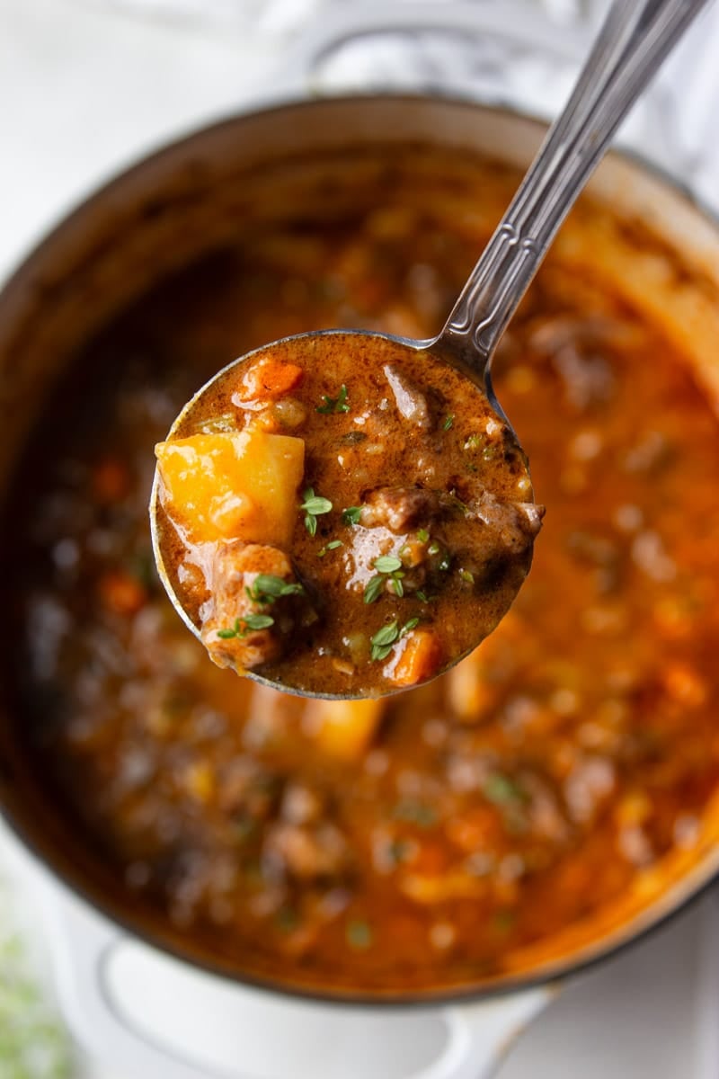 Oxtail soup on a spoon, a pot of oxtail soup, fresh thyme and a white and black dish towel. 