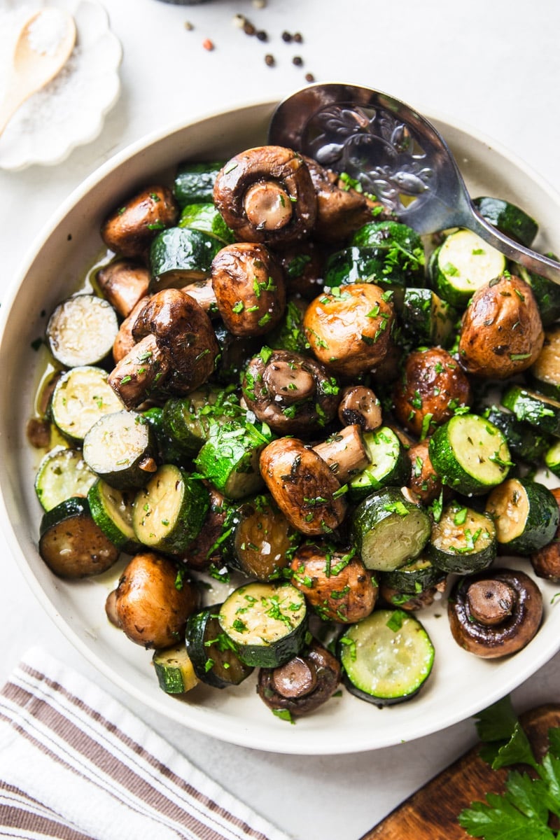 Pan sauteed zucchini and mushrooms with a silver spoon and parsley garnish, white and brown dish towel. 