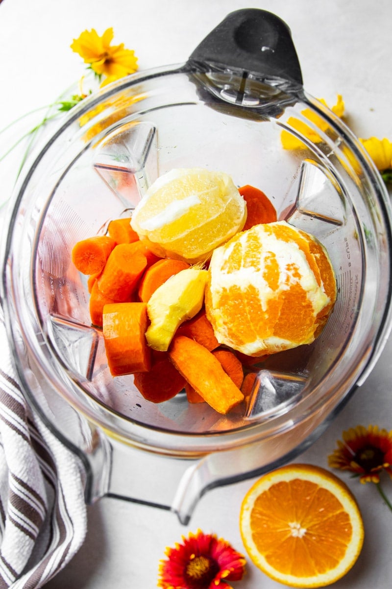Peel orange, carrots and ginger in a blender, orange halves, flowers, and brown and white dish towel. 