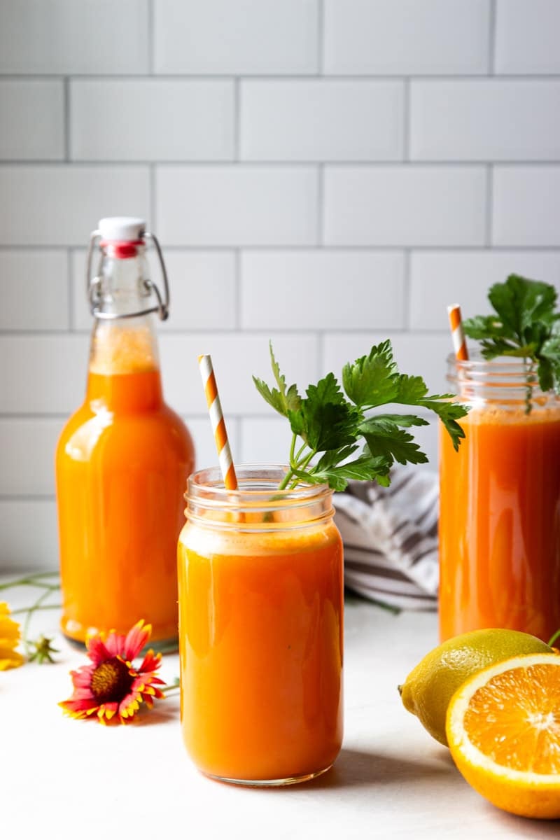 Vitamin C anti inflammatory juice in glass jars with a straw and parsley, flowers, lemons, oranges, white and brown dish towel.
