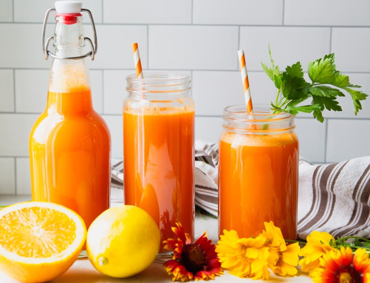 Vitamin C anti inflammatory juice in glass jars with a straw and parsley, flowers, lemons, oranges, white and brown dish towel.