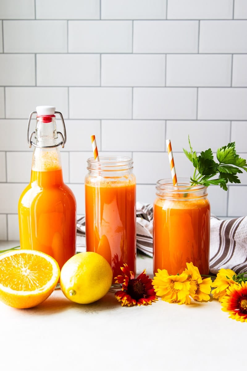 Vitamin C anti inflammatory juice in glass jars with a straw, flowers, lemons, oranges, white and brown dish towel.