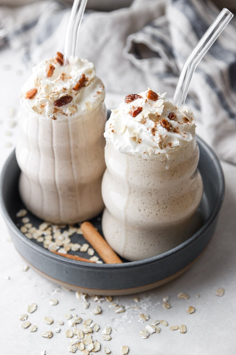 Apple pie protein smoothies with glass straws, oats, cinnamon stick, and white and blue dish towel.