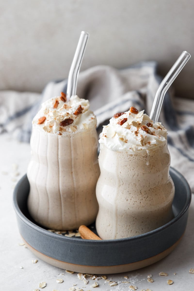 Apple pie protein smoothies with glass straws, cinnamon sticks, oats, blue and white dish towel.