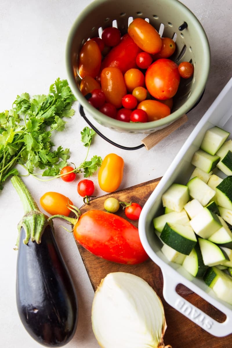 A variety of tomatoes, cilantro, eggplant, onion, cubed zucchini in a white casserole dish, and a wooden cutting board.