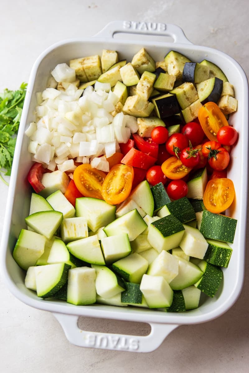 Chopped onions, tomatoes, zucchini and eggplant in a white casserole dish. 