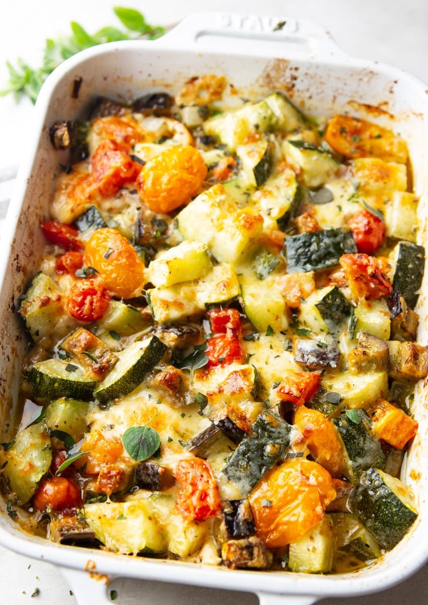 Baked zucchini casserole in a white baking dish, grey and white dish towel, and oregano. 