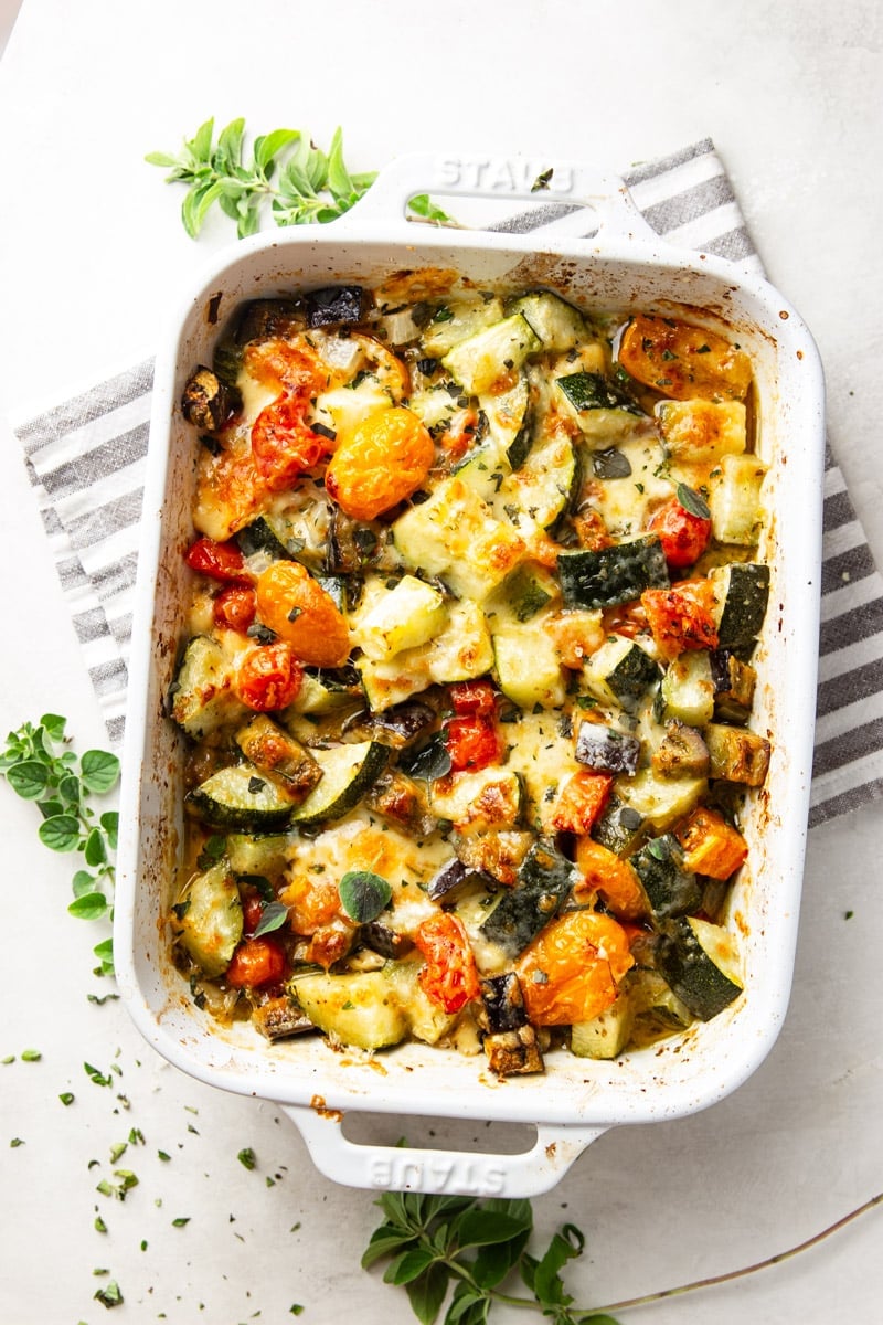 Baked zucchini casserole in a white baking dish, grey and white dish towel, and oregano. 