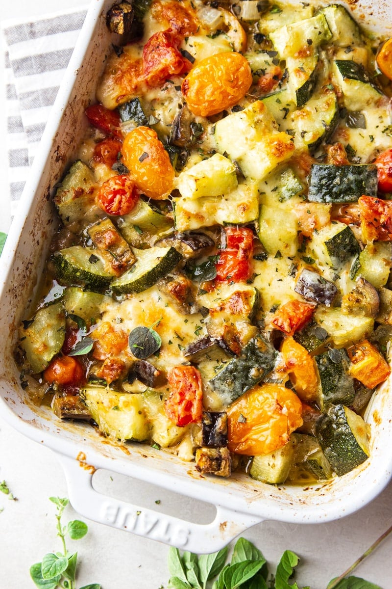 Baked zucchini casserole in a white baking dish with a white and grey dish towel and oregano. 